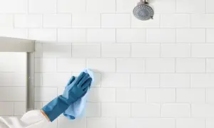 Gloved hand wipes white shower tiles, demonstrating how to clean shower tiles for a sparkling finish.