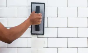 A hand applies grout to white subway tiles. The difference between sanded grout vs unsanded grout is visible in the texture.