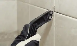 Gloved hand uses a grout removal tool to scrape away old grout, preparing for new grout over old grout.
