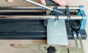Hands guide a scoring wheel on a tile cutter, demonstrating how to cut ceramic tile by hand for a precise DIY project.