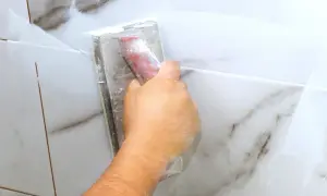 A hand uses a grout float to apply white grout to marble tiles. The grout is still wet, so it's too early to know how long does it take for grout to set.