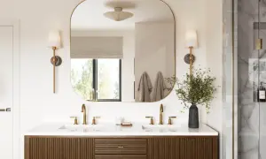 A modern bathroom vanity with dual sinks and brass fixtures. The large, rounded mirror reflects the window and wall sconces, effectively using mirrors to reflect light and brighten the space.