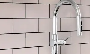 A modern chrome faucet stands against a backdrop of light pink subway tiles with smooth grout.