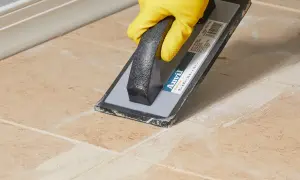 Yellow-gloved hand uses a grout float to remove old grout from tan tiles, demonstrating how to remove grout from tile.