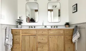A beautiful DIY bathroom vanity with a natural wood finish, double sinks, and brass fixtures. Two framed mirrors hang above, reflecting the bright, airy space.