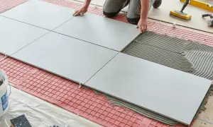 Tiling floors in a kitchen with large grey tiles and adhesive. Tools and a bucket of grout are nearby.