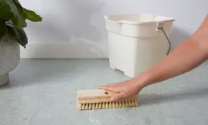 A hand scrubs a linoleum floor with a brush, with a bucket nearby. This shows how to clean linoleum floors effectively.