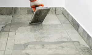 Gloved hand applies grout to tiled floor. Wondering how long for grout to dry? This process is key to a finished look.