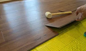 A hand is installing a dark wood-look LVT plank flooring over yellow underlayment. A rubber mallet rests nearby.
