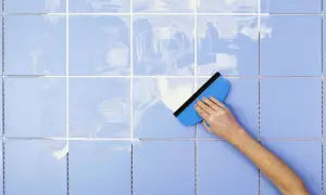 A hand uses a blue squeegee to spread white grout on light blue tiles, demonstrating how to clean tile grout.