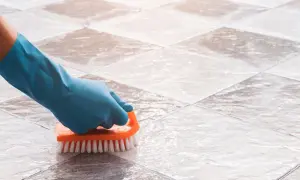 Gloved hand scrubs tile grout with an orange brush, demonstrating how to clean grout on a tile floor.