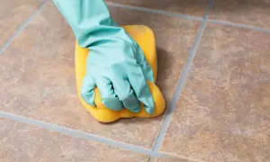 A gloved hand scrubs unglazed ceramic tile with a yellow sponge, demonstrating how to clean the porous surface.