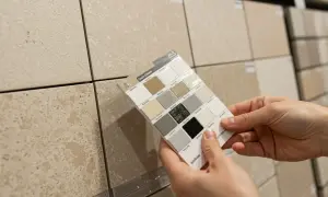 Hands hold a grout color swatch against beige tiles, exploring options for the best grout color for beige tile.