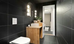 A modern bathroom with black tiles. Natural wood vanity, white sink, and toilet complement the dark tiles, showing what colour goes with black tiles in bathroom.