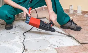 A person in green overalls uses a jackhammer to remove ceramic tile flooring, breaking up the old tiles and concrete.