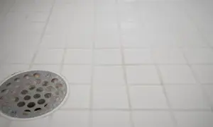 White tiled shower floor with a metal drain cover. The grout lines show signs of wear, indicating a need for tile grout repair.