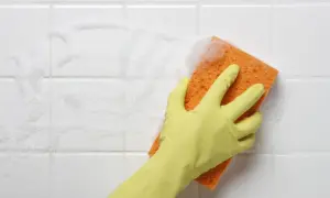 A gloved hand scrubs white bathroom tiles with a soapy orange sponge, showing the best way to clean tiles.