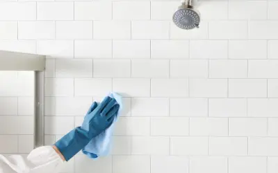 Gloved hand wipes white shower tiles, demonstrating how to clean shower tiles for a sparkling finish.