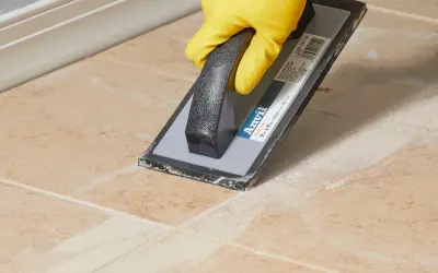 Yellow-gloved hand uses a grout float to remove old grout from tan tiles, demonstrating how to remove grout from tile.