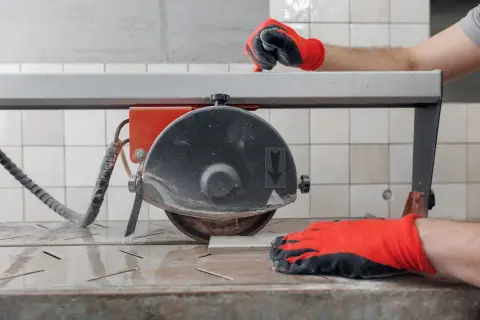 Hands in red gloves guide a tile on a wet saw, demonstrating how to use a wet saw for precise cuts.