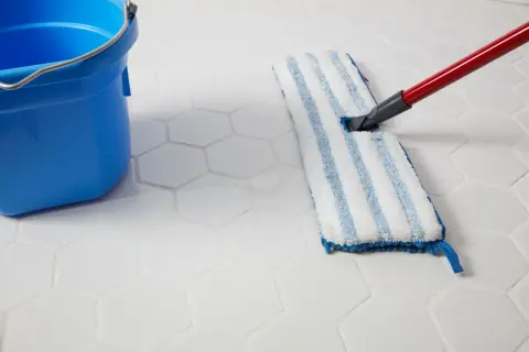 A blue bucket and a mop are used to clean white hexagonal porcelain tile floors.