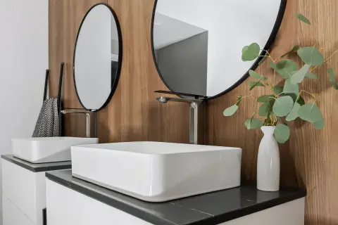 Modern bathroom with two white vessel sinks, sleek faucets, round mirrors, and a eucalyptus branch in a vase.