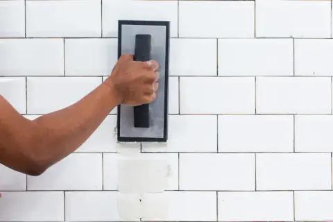 A hand applies grout to white subway tiles. The difference between sanded grout vs unsanded grout is visible in the texture.