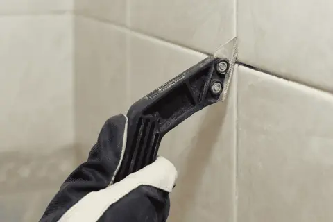 Gloved hand uses a grout removal tool to scrape away old grout, preparing for new grout over old grout.