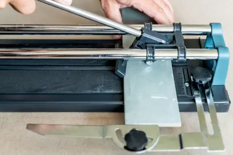 Hands guide a scoring wheel on a tile cutter, demonstrating how to cut ceramic tile by hand for a precise DIY project.