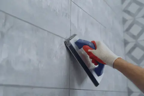 Hand in glove using a trowel for repairing grouting between light grey tiles.