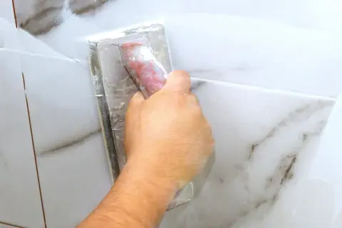 A hand uses a grout float to apply white grout to marble tiles. The grout is still wet, so it's too early to know how long does it take for grout to set.