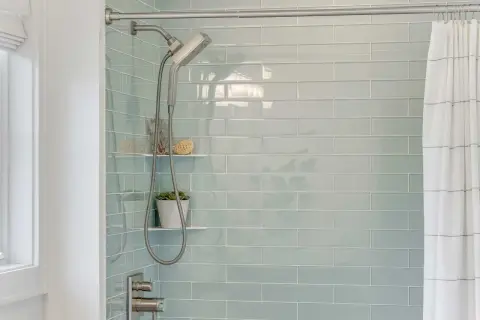 Modern shower with light blue subway tiles. A shower head and hose hang next to a small plant. This is the best way to regrout tiles for a fresh look.