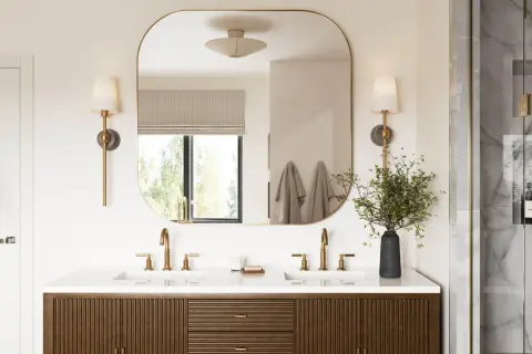 A modern bathroom vanity with dual sinks and brass fixtures. The large, rounded mirror reflects the window and wall sconces, effectively using mirrors to reflect light and brighten the space.