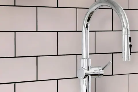 A modern chrome faucet stands against a backdrop of light pink subway tiles with smooth grout.