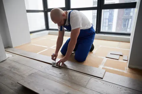 A man in blue overalls measures a laminate plank, demonstrating how to install laminate flooring.
