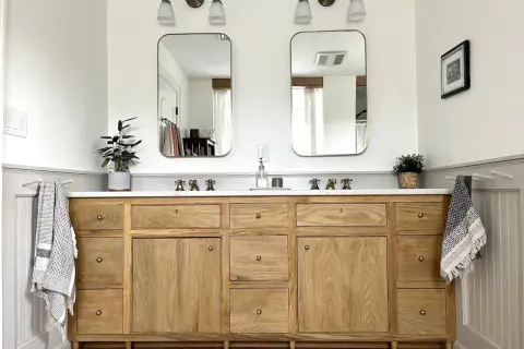 A beautiful DIY bathroom vanity with a natural wood finish, double sinks, and brass fixtures. Two framed mirrors hang above, reflecting the bright, airy space.