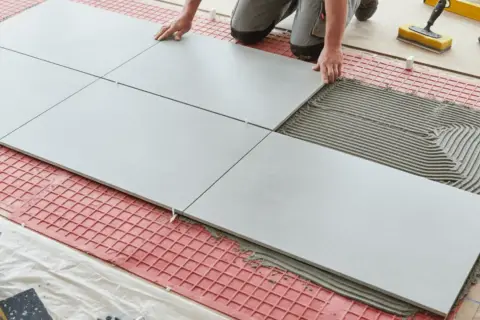Tiling floors in a kitchen with large grey tiles and adhesive. Tools and a bucket of grout are nearby.