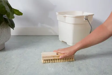 A hand scrubs a linoleum floor with a brush, with a bucket nearby. This shows how to clean linoleum floors effectively.