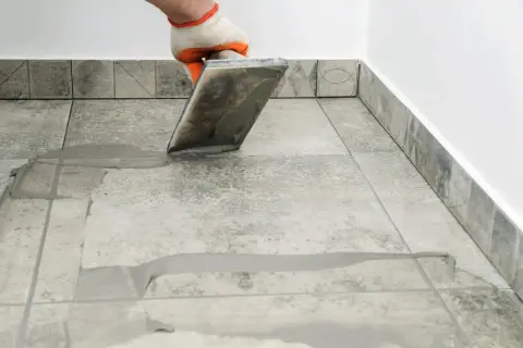 Gloved hand applies grout to tiled floor. Wondering how long for grout to dry? This process is key to a finished look.