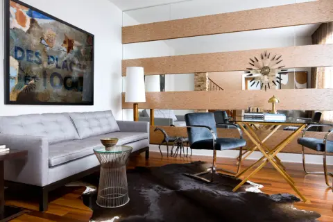 A large wall mirror with wood accents offers a stylish way to decorate around it. A grey sofa, abstract art, and a glass desk complete the modern living space.