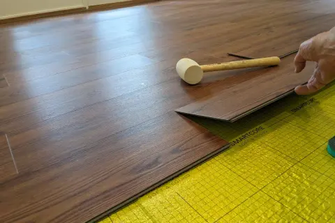A hand is installing a dark wood-look LVT plank flooring over yellow underlayment. A rubber mallet rests nearby.