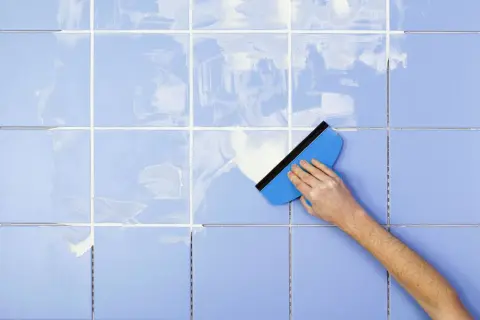 A hand uses a blue squeegee to spread white grout on light blue tiles, demonstrating how to clean tile grout.