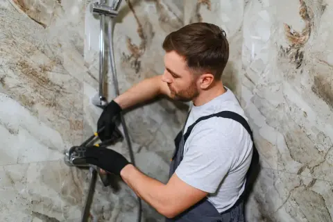 A plumber in black gloves works on a new shower install, tightening a fixture against a marble-patterned wall.