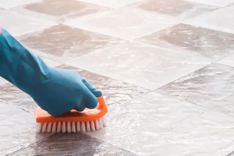 Gloved hand scrubs tile grout with an orange brush, demonstrating how to clean grout on a tile floor.