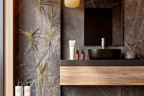 Modern bathroom vanity ideas featuring a floating wood shelf, dark vessel sink, and marble walls. A tall plant adds a natural touch.