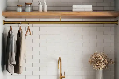 A laundry room with white tile beige grout walls, a wooden shelf, and a brass rod holding clothes.