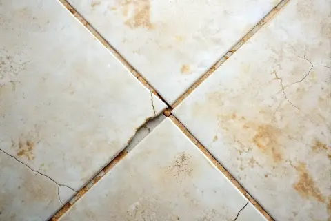 Cracked grout between beige floor tiles. Consider how often should you regrout floor tiles to prevent this damage and maintain your floor's integrity.