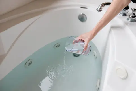 A hand pours liquid from a glass into a jetted bathtub, part of a process for how to clean bathtub.