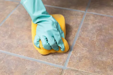 A gloved hand scrubs unglazed ceramic tile with a yellow sponge, demonstrating how to clean the porous surface.