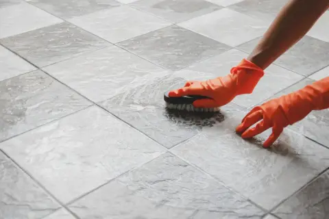 Hands in orange gloves scrub a tiled floor. The person is working to polish porcelain tile, making it shine.